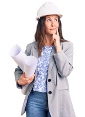 Young beautiful brunette woman wearing architect hardhat holding blueprint serious face thinking about question with hand on chin, thoughtful about confusing idea