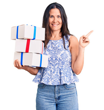 Young beautiful brunette woman holding birthday gift smiling happy pointing with hand and finger to the side