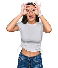 Young hispanic girl wearing casual white t shirt doing ok gesture like binoculars sticking tongue out, eyes looking through fingers. crazy expression.