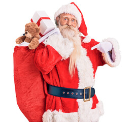 Old senior man with grey hair and long beard wearing santa claus costume holding bag with presents pointing finger to one self smiling happy and proud