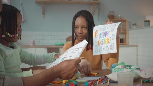 African American teenage girl with father and little sister making Easter crafts and decorations and coloring eggs while sitting together at table in cozy kitchen