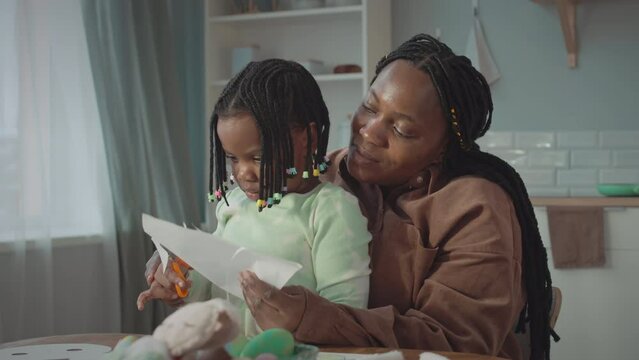 Happy Black woman with little daughter sitting on her laps making crafts and decorations for Easter together at cozy home