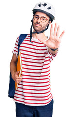 Young hispanic man wearing student backpack and bike helmet holding binder with open hand doing stop sign with serious and confident expression, defense gesture