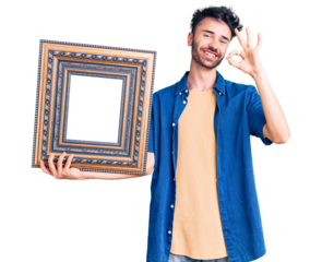 Young hispanic man holding empty frame doing ok sign with fingers, smiling friendly gesturing excellent symbol