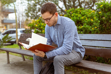 Hombre joven, caucásico, de 25 años, leyendo un libro, en un banco de un parque.