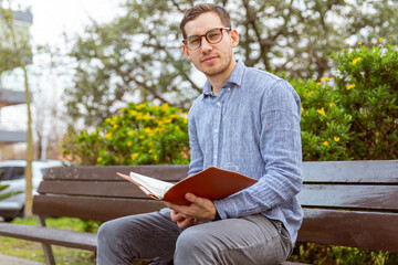 Hombre joven, caucásico, de 25 años, leyendo un libro, en un banco de un parque.
