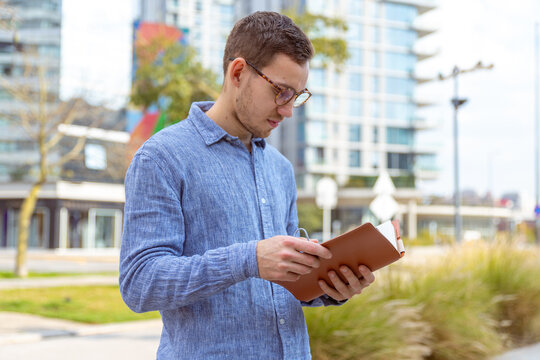 Hombre joven, cauc&aacute;sico, de 25 a&ntilde;os, con lentes, leyendo un libro, al aire libre.