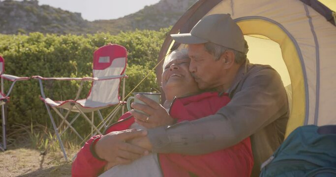 Happy senior biracial couple sitting at tent in mountains and embracing on sunny day, in slow motion