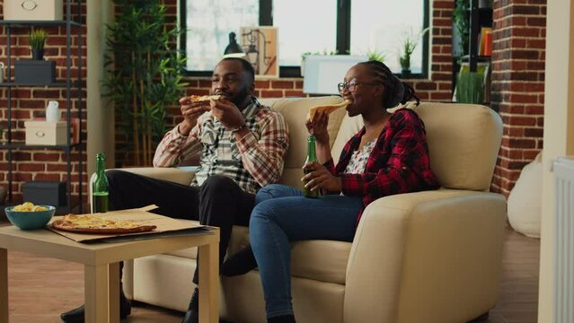 African american people eating slices of pizza in front of tv, binge watching favorite show at home. Relaxed modern couple serving fast food meal and drinking bottles of beer.