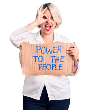 Young blonde plus size woman holding power to the people banner smiling happy doing ok sign with hand on eye looking through fingers