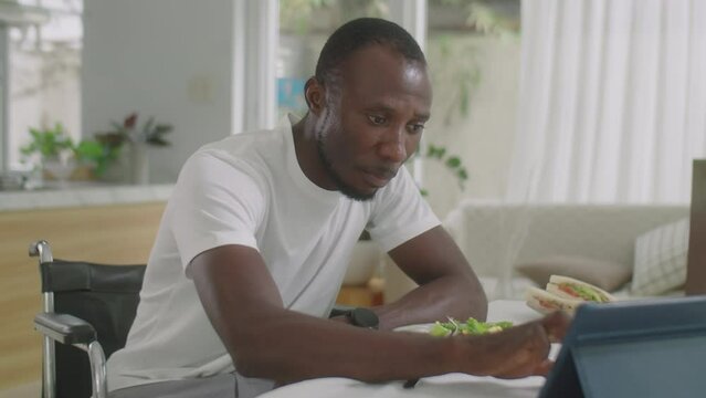 African American man in wheelchair eating salad, watching something on digital tablet and smiling while having dinner at home