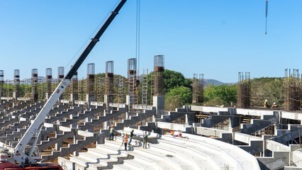 A group of Latino workers is installing concrete steps for a stadium with the help of a crane. The workers wear helmets and safety vests as they place the steps in place. The stadium has a metal © Carlos