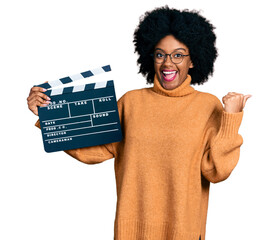 Young african american woman holding video film clapboard pointing thumb up to the side smiling...