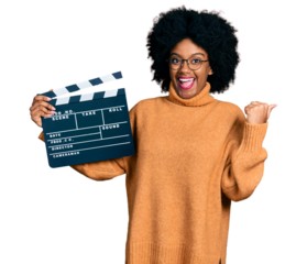 Young african american woman holding video film clapboard pointing thumb up to the side smiling happy with open mouth