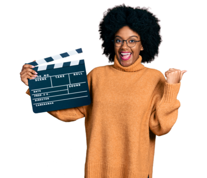 Young african american woman holding video film clapboard pointing thumb up to the side smiling happy with open mouth