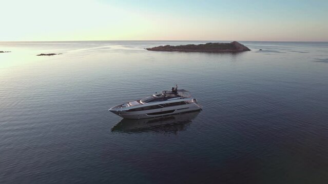 Aerial orbit view of a wealthy billionaire luxury Riva Corsaro super motor yacht staying at anchor in mediterranean sea during colorful sunrise with green islands in the beautiful bay