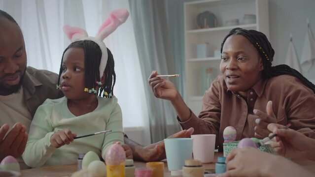 Happy African American family of four coloring Easter eggs together sitting at wooden table in bright cozy kitchen