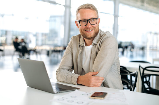 Photo of a successful caucasian man with glasses, in stylish casual wear, IT specialist, company seo, programmer, digital developer, sits at a desk with a laptop in modern office, looking away, smiles