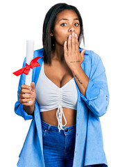 Beautiful hispanic woman holding graduate degree diploma covering mouth with hand, shocked and...