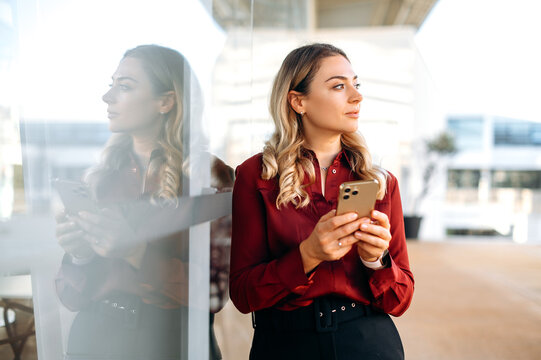 Lovely attractive caucasian woman stands outdoors, uses her smartphone, writes a message, texts with friends in social media, searches for information, looks away, thinking about message