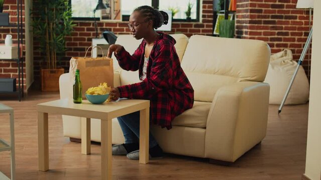 Young adult taking out hamburger and fries, eating fast food takeaway meal from delivery bag on couch. Woman enjoying dinner wth burger and snacks, watching comedy movie on television.