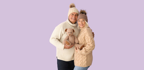 Happy mature couple in winter clothes and with toy on lilac background