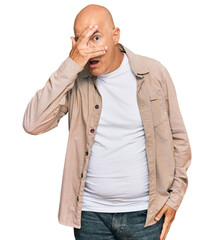 Middle age bald man wearing casual clothes peeking in shock covering face and eyes with hand, looking through fingers with embarrassed expression.