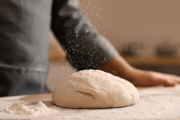 Male baker sprinkling dough with flour at table in kitchen, closeup