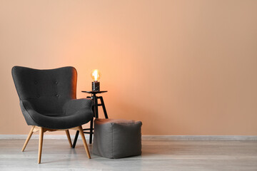 Stylish grey armchair, pouf and glowing lamp near beige wall