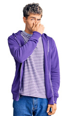 Young handsome man wearing casual purple sweatshirt smelling something stinky and disgusting,...