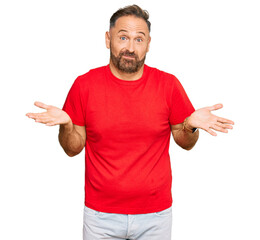 Handsome middle age man wearing casual red tshirt clueless and confused expression with arms and hands raised. doubt concept.