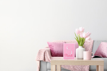 Vase with tulips, cup and greeting card for Women's Day on table in living room