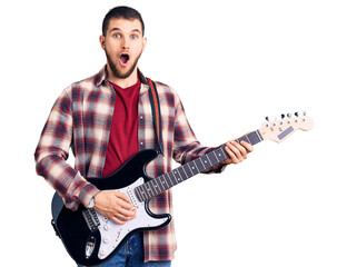 Young handsome man playing electric guitar scared and amazed with open mouth for surprise,...