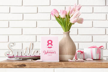 Vase with tulips, gifts, magazines and greeting card for Women's Day on table near white brick wall