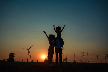 Fototapeta na wymiar Silhouettes of Happy family father, mother and child daughter sits on the shoulders of his father with Wind turbine farm power generator in beautiful nature landscape for production of renewable green