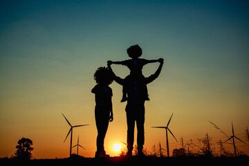 Silhouettes of Happy family father, mother and child daughter sits on the shoulders of his father with windmills for electricity generation at sunrise by producing sustainable energy 