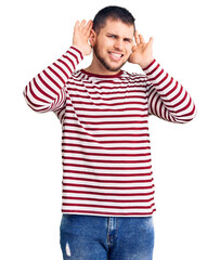 Young handsome man wearing striped sweater trying to hear both hands on ear gesture, curious for gossip. hearing problem, deaf