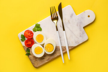 Board with delicious boiled eggs, tomatoes, cutlery and parsley on yellow background