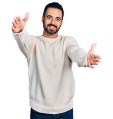 Naklejka premium Young hispanic man with beard wearing casual white sweater looking at the camera smiling with open arms for hug. cheerful expression embracing happiness.