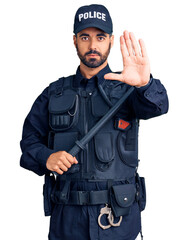 Young hispanic man wearing police uniform holding baton with open hand doing stop sign with serious and confident expression, defense gesture