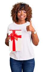 Young african american woman holding gift smiling happy and positive, thumb up doing excellent and approval sign