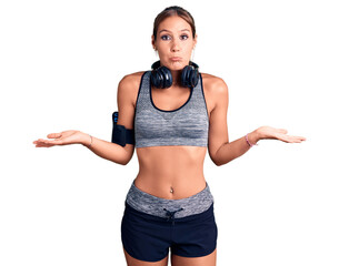 Young beautiful hispanic woman wearing gym clothes and using headphones clueless and confused...