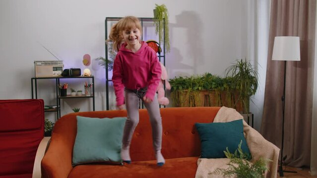 Funny young blonde child kid girl dancing trendy social media dance while listening to the music at home in cozy living room. Female little teen toddler having fun, jumping, playing game on sofa alone