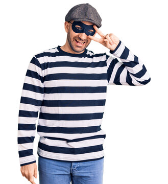 Young handsome man wearing burglar mask doing peace symbol with fingers over face, smiling cheerful showing victory