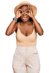 Young black woman wearing summer hat doing ok gesture like binoculars sticking tongue out, eyes looking through fingers. crazy expression.