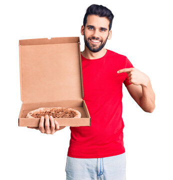 Young handsome man with beard holding delivery cardoboard with italian pizza pointing finger to one self smiling happy and proud