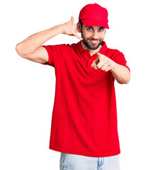 Young handsome man with beard wearing delivery uniform smiling doing talking on the telephone gesture and pointing to you. call me.