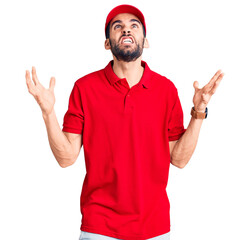 Naklejka premium Young handsome man with beard wearing delivery uniform crazy and mad shouting and yelling with aggressive expression and arms raised. frustration concept.
