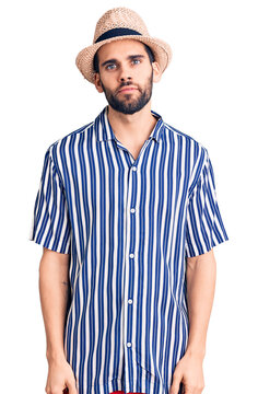 Young handsome man with beard wearing summer hat and striped shirt relaxed with serious expression on face. simple and natural looking at the camera.