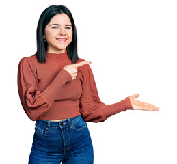 Young brunette woman with blue eyes wearing elegant sweater amazed and smiling to the camera while presenting with hand and pointing with finger.